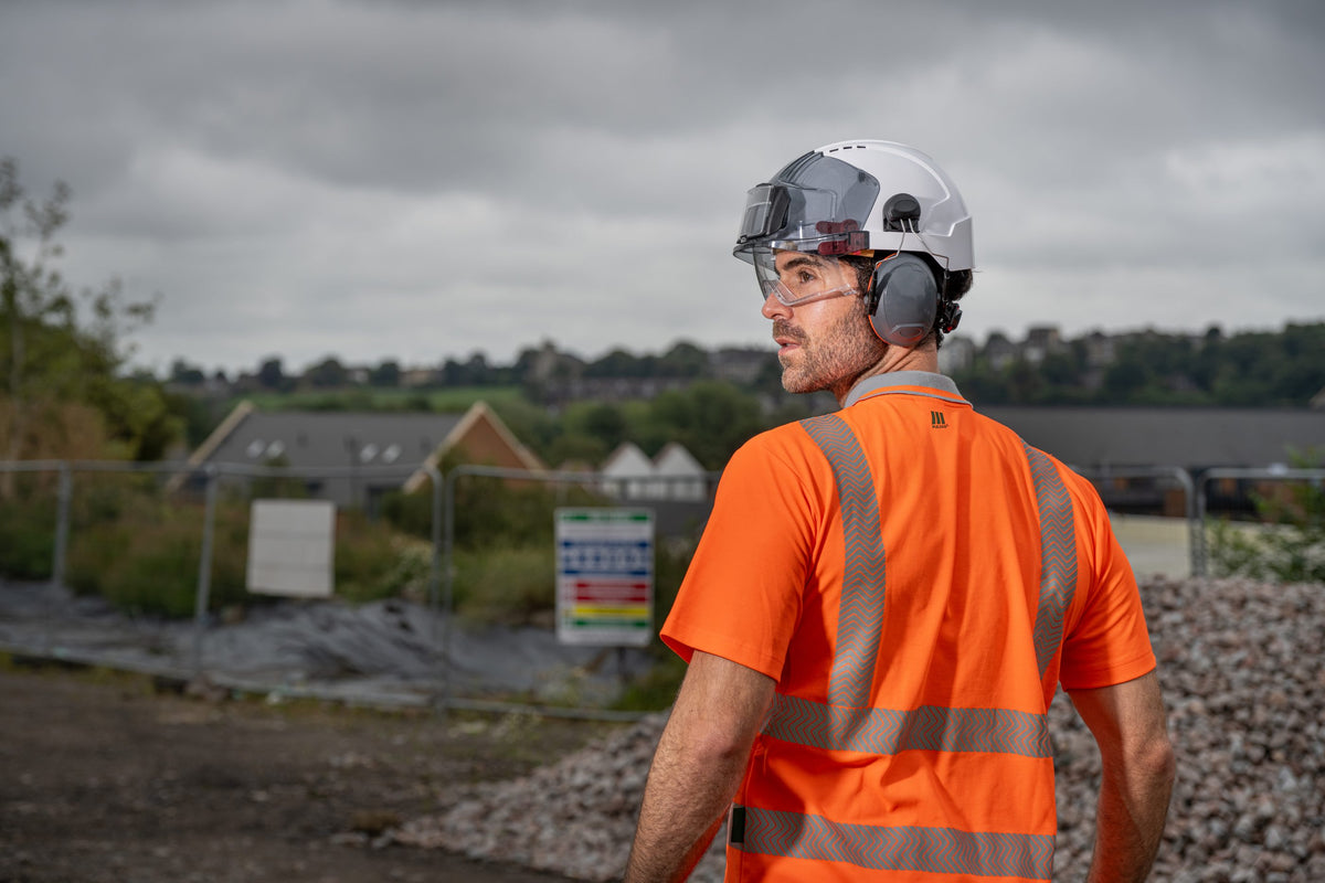 Site worker in a PULSAR orange hi-vis polo shirt and white safety helmet with ear defenders, looking out across a construction site
