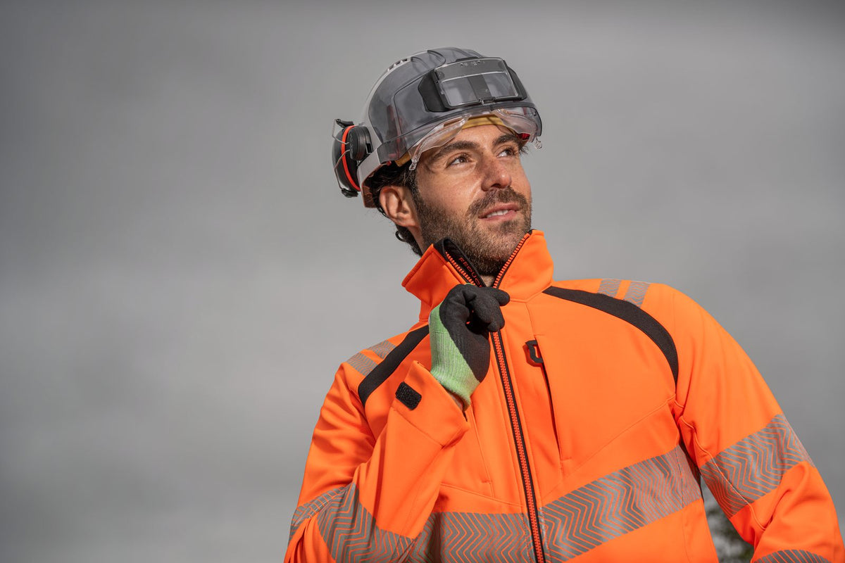 Rail worker in an orange hi-vis softshell jacket, safety helmet with visor and ear defenders, and work gloves - railway workwear and PPE from The Workwear Co
