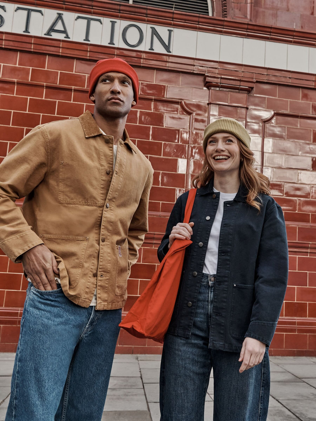 Two people in Stanley/Stella jackets and beanies outside a red-brick station